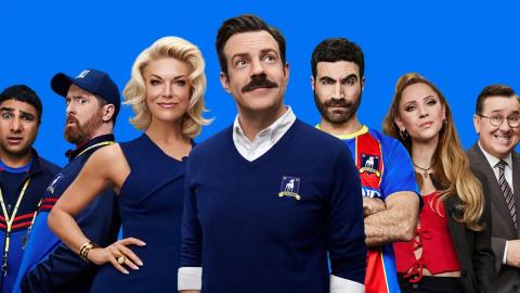 The cast of the TV show Ted Lasso face the camera against a blue background. Characters pictured from left to right are Nate, Coach Beard, Rebecca, Ted, Roy, Keeley, and Higgins.