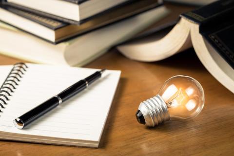 A pen rests on an open notebook next to an illuminated lightbulb.