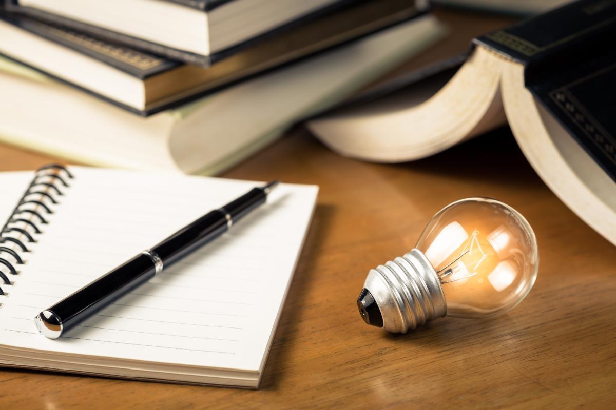 A pen rests on an open notebook next to an illuminated lightbulb.